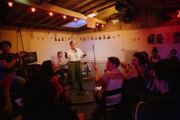 Jo Chiang performing on stage in a cozy room filled with happy people.
