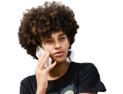 Young person with curly haired styled in an afro. They are wearing a black t-shirt and are holding a smart phone to their ear as if they are talking on the phone.