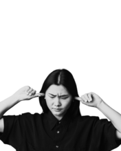 Young woman with black hair and a black shirt has a finger in each of her ears to try and block out sound. Her eyes are closed and she looks overwhelmed.